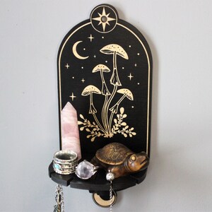 May include: A black wooden shelf with a crescent moon and stars design. The shelf has a gold painted mushroom design and a small turtle figurine. The shelf is decorated with a pink crystal, rings, and a necklace.