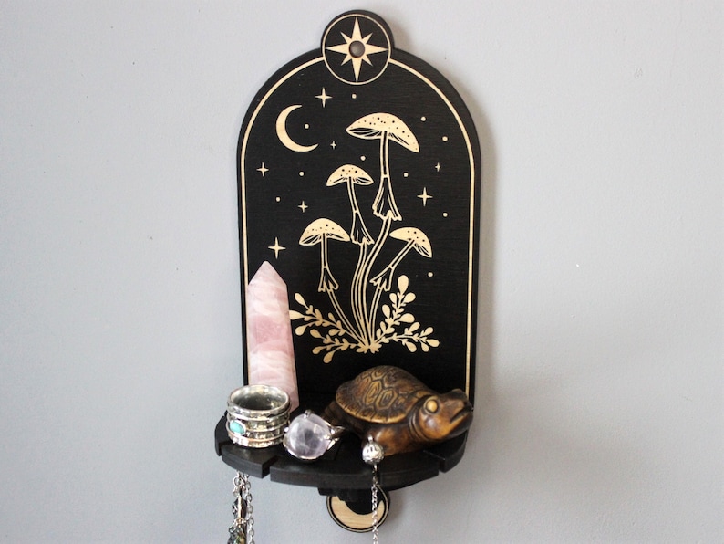 May include: A black wooden shelf with a crescent moon and stars design. The shelf has a gold painted mushroom design and a small turtle figurine. The shelf is decorated with a pink crystal, rings, and a necklace.
