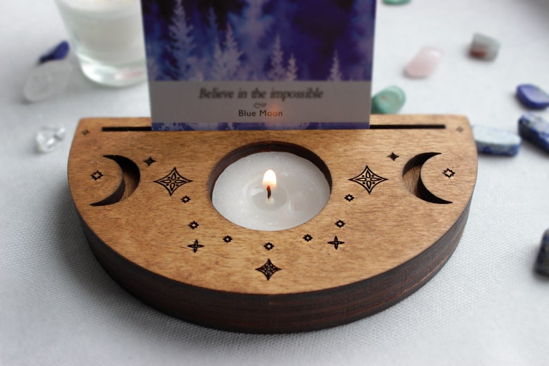 May include: Wooden candle holder with a crescent moon design and a small candle burning inside. The holder has a slot for a card that reads "Believe in the impossible of Blue Moon".