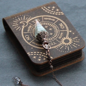 May include: A wooden box with a pendulum inside. The box has a circular design with the words "Yes", "No", "Maybe", and "Rephrase" printed on it. The pendulum is made of a clear crystal with a metal top.