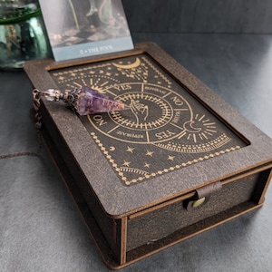 May include: A dark brown wooden divination box with a hinged lid and clasp. The lid is engraved with a circular design featuring the words "YES", "NO", and "REPHRASE". A purple crystal pendulum hangs over the box. A tarot card is in the background.