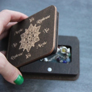 May include: A wooden divination box with a hinged lid, featuring engraved text and a central floral design. The lid is open, revealing a compartment with a beaded necklace inside. The box is brown.
