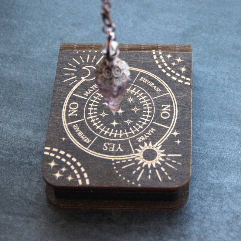 Puede incluir: Un tablero de p&eacute;ndulo de madera con un gr&aacute;fico circular que presenta las palabras "S&iacute;", "No", "Tal vez" y "Reformular". Un p&eacute;ndulo de cristal morado cuelga sobre el tablero.