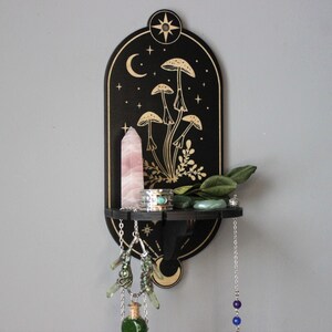 May include: Black wooden shelf with a crescent moon and stars design. The shelf features a gold mushroom illustration and a small shelf for displaying crystals and other items.