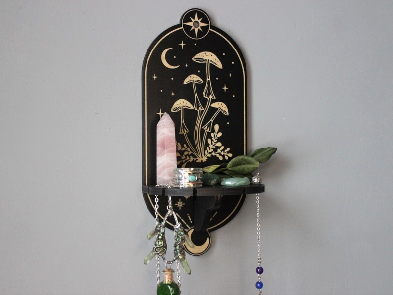 May include: Black wooden shelf with a crescent moon and stars design. The shelf features a gold mushroom illustration and a small shelf for displaying crystals and other items.