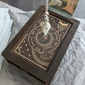 May include: A wooden divination box with a pendulum and tarot cards. The box is dark brown with gold-colored etched designs, including a hand, stars, and the words "YES", "NO", and "MAYBE". The pendulum is silver-toned with a pear-shaped crystal.
