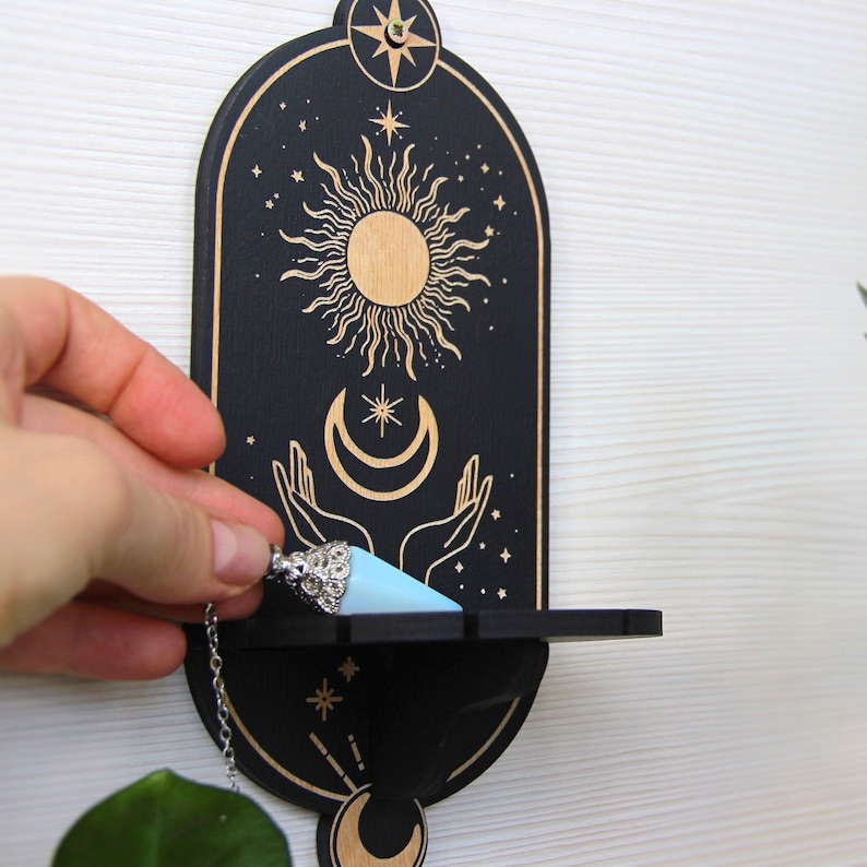 May include: Black wooden wall decor with a shelf, featuring a gold sun, moon, and stars design. A blue crystal pendulum hangs from the shelf.