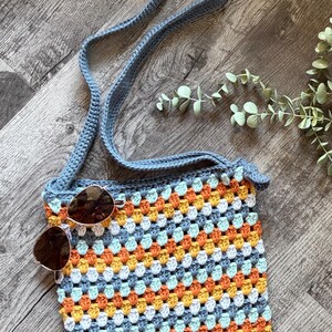 Puede incluir: Un bolso bandolera de ganchillo con un patr&oacute;n de cuadrados de la abuela multicolor en tonos naranja, amarillo, blanco y azul claro, con una correa y borde en azul s&oacute;lido. Unas gafas de sol tintadas en marr&oacute;n con montura dorada descansan sobre el bolso.