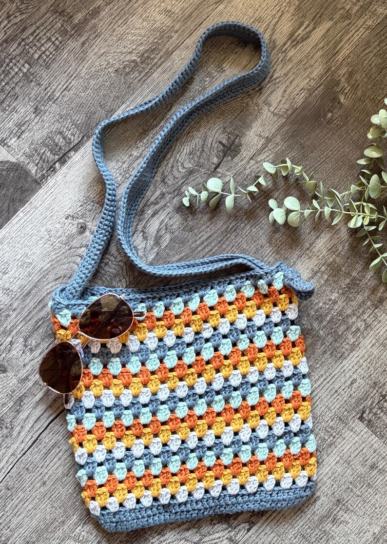 Puede incluir: Un bolso bandolera de ganchillo con un patr&oacute;n de cuadrados de la abuela multicolor en tonos naranja, amarillo, blanco y azul claro, con una correa y borde en azul s&oacute;lido. Unas gafas de sol tintadas en marr&oacute;n con montura dorada descansan sobre el bolso.