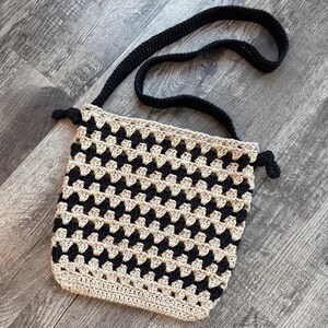Puede incluir: Un bolso bandolera de ganchillo con un patr&oacute;n repetitivo de cuadrados granny en negro y beige. El bolso tiene una correa de hombro negra lisa y detalles laterales anudados en negro. La parte inferior est&aacute; rematada con una fila de puntos beige.