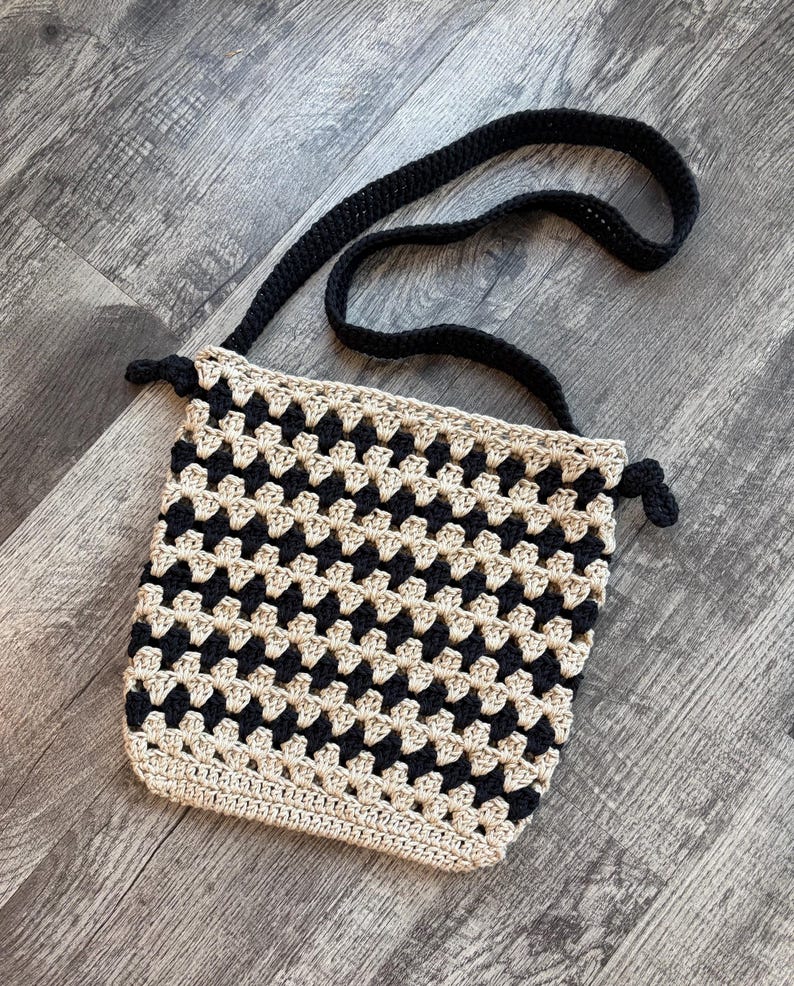 Puede incluir: Un bolso bandolera de ganchillo con un patr&oacute;n repetitivo de cuadrados granny en negro y beige. El bolso tiene una correa de hombro negra lisa y detalles laterales anudados en negro. La parte inferior est&aacute; rematada con una fila de puntos beige.