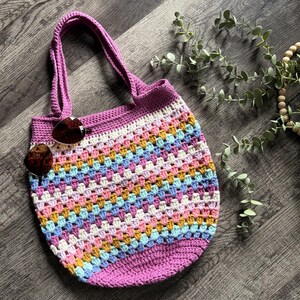 Puede incluir: Un bolso tote de ganchillo con un patr&oacute;n de puntos granny multicolor en tonos rosa, amarillo, azul y blanco, con una base morada s&oacute;lida y asas a juego. Un par de gafas de sol con cristales marrones descansa en la parte superior izquierda.