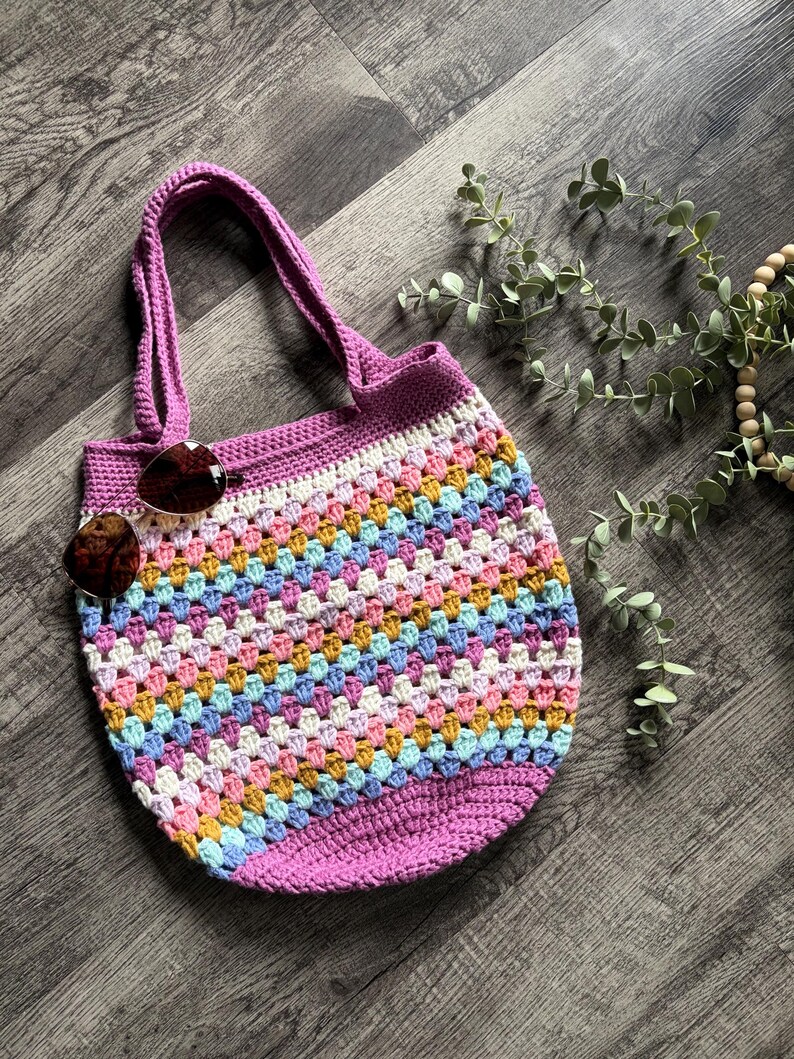 Puede incluir: Un bolso tote de ganchillo con un patr&oacute;n de puntos granny multicolor en tonos rosa, amarillo, azul y blanco, con una base morada s&oacute;lida y asas a juego. Un par de gafas de sol con cristales marrones descansa en la parte superior izquierda.
