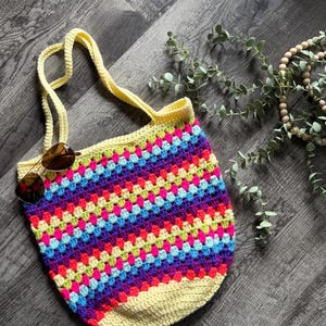 Puede incluir: Un bolso tote de ganchillo con un vibrante patr&oacute;n de cuadrados de la abuela multicolor en tonos rosa, morado, azul y verde lima, con una base de color amarillo p&aacute;lido s&oacute;lido y asas amarillas a juego. Unas gafas de sol tintadas en marr&oacute;n descansan sobre el bolso.