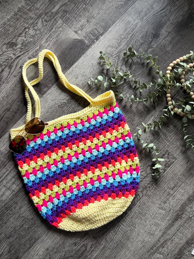 Puede incluir: Un bolso tote de ganchillo con un vibrante patr&oacute;n de cuadrados de la abuela multicolor en tonos rosa, morado, azul y verde lima, con una base de color amarillo p&aacute;lido s&oacute;lido y asas amarillas a juego. Unas gafas de sol tintadas en marr&oacute;n descansan sobre el bolso.
