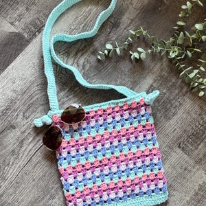 Puede incluir: Un bolso bandolera de ganchillo con un patr&oacute;n de cuadrados de la abuela multicolor en tonos rosa, azul y violeta claro, acabado con un borde verde menta s&oacute;lido y una correa a juego. Un par de gafas de sol marrones descansan sobre el bolso.