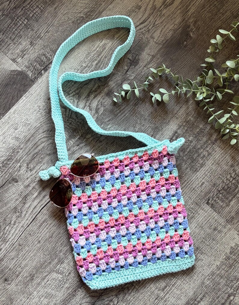 Puede incluir: Un bolso bandolera de ganchillo con un patr&oacute;n de cuadrados de la abuela multicolor en tonos rosa, azul y violeta claro, acabado con un borde verde menta s&oacute;lido y una correa a juego. Un par de gafas de sol marrones descansan sobre el bolso.