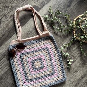 Puede incluir: Un bolso de mano tejido a ganchillo hecho a mano con un motivo cuadrado de abuela en tonos alternos de rosa claro, azul suave y gris. El bolso tiene dos asas integradas en hilo rosa claro s&oacute;lido y un par de gafas de sol de color marr&oacute;n oscuro.