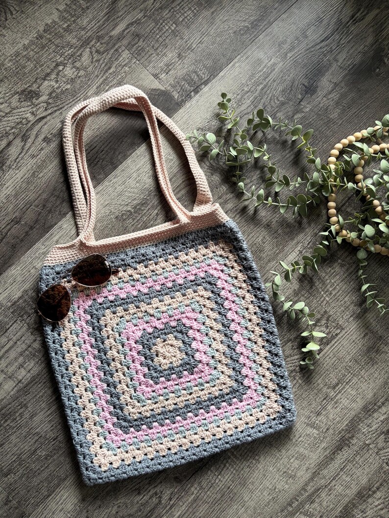 Puede incluir: Un bolso de mano tejido a ganchillo hecho a mano con un motivo cuadrado de abuela en tonos alternos de rosa claro, azul suave y gris. El bolso tiene dos asas integradas en hilo rosa claro s&oacute;lido y un par de gafas de sol de color marr&oacute;n oscuro.