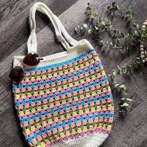 Puede incluir: Un bolso de ganchillo con un patr&oacute;n multicolor de puntos de lana rosa, amarilla, azul y verde. El bolso tiene un borde superior blanco liso con dos asas a juego y una base blanca lisa. Unas gafas de sol marrones descansan sobre el bolso.