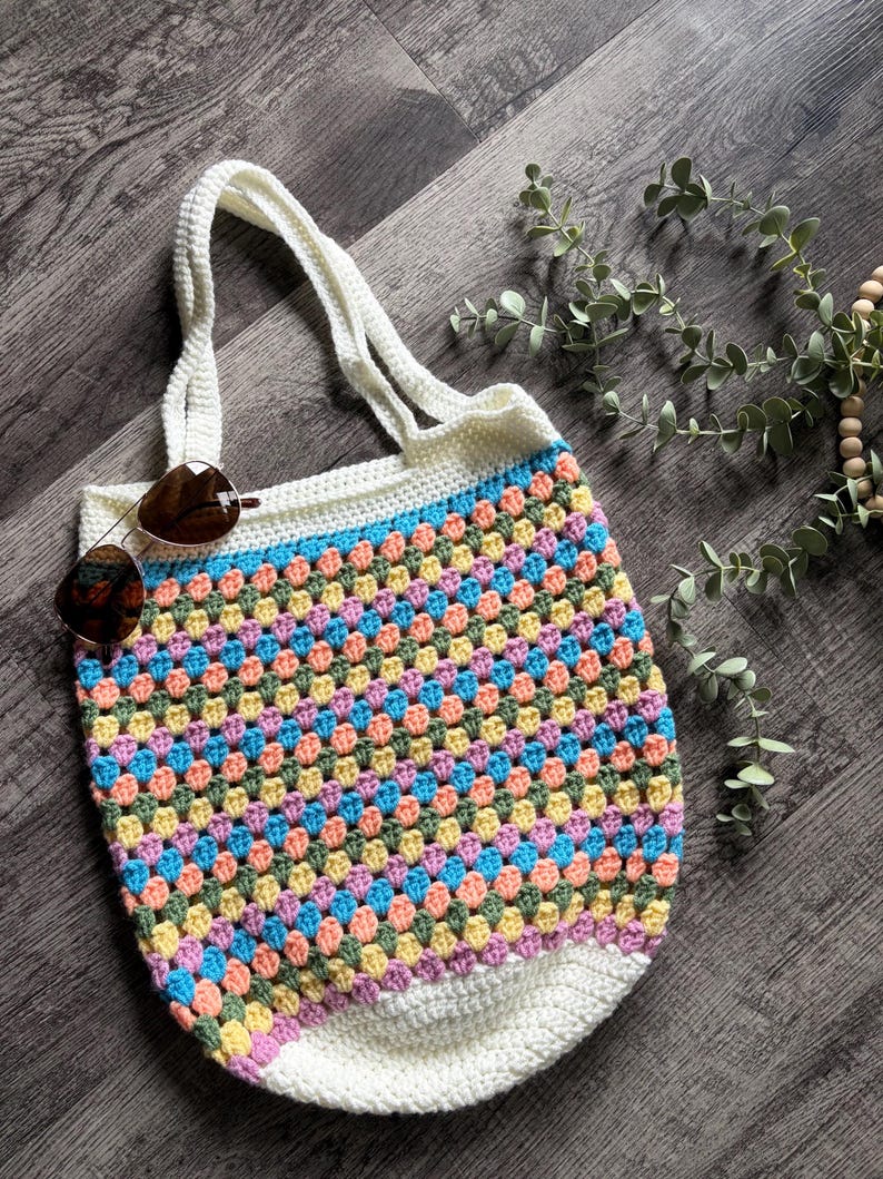 Puede incluir: Un bolso de ganchillo con un patr&oacute;n multicolor de puntos de lana rosa, amarilla, azul y verde. El bolso tiene un borde superior blanco liso con dos asas a juego y una base blanca lisa. Unas gafas de sol marrones descansan sobre el bolso.