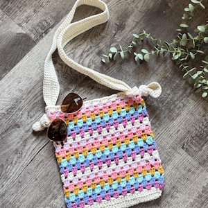 Puede incluir: Un bolso rectangular de ganchillo con un patr&oacute;n de cuadrados de la abuela multicolor en tonos rosa, amarillo, azul y blanco. El bolso tiene una correa larga de ganchillo color crema y unas gafas de sol tintadas en marr&oacute;n sobre el frente.