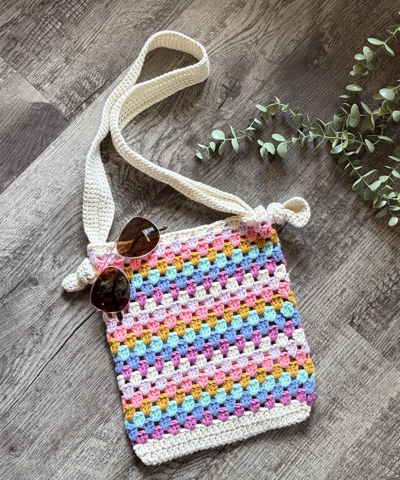 Puede incluir: Un bolso rectangular de ganchillo con un patr&oacute;n de cuadrados de la abuela multicolor en tonos rosa, amarillo, azul y blanco. El bolso tiene una correa larga de ganchillo color crema y unas gafas de sol tintadas en marr&oacute;n sobre el frente.