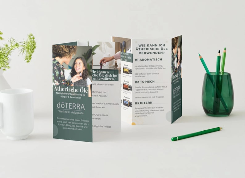 May include: A product display featuring a brochure about essential oils, with the dōTERRA logo. The brochure is open, revealing text and images. A green glass vase holds green pencils, and a white mug is on the left.