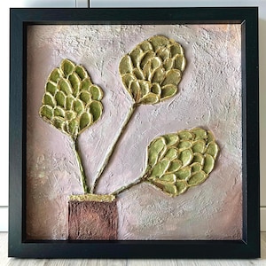 May include: A square framed wall art piece featuring a textured, light pink and beige background. Three stylized, raised green plant forms with gold-toned edges emerge from a rectangular, dark brown pot at the bottom center of the composition.