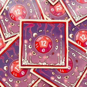 Pu&ograve; includere: Un adesivo illustrato viola e rosso con un dado a 12 facce con candele luminose e occhi che lo circondano. Il dado &egrave; rosso con numeri bianchi e il testo "12" &egrave; visibile sul dado.
