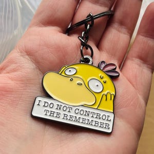 May include: A yellow and black keychain with a cartoon character and the text "I DO NOT CONTROL THE REMEMBER". The keychain has a black metal ring and a yellow duck-like character with white eyes and a large yellow beak.