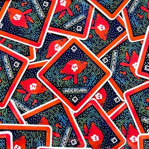 May include: A stack of square stickers with a black background and a red border. The stickers feature a mushroom with a skull on top, surrounded by a swirling pattern of green, blue, and white lines. The text "UNDERDARK" is printed in white on the stickers.