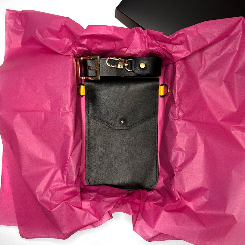 May include: A black leather phone case with a yellow strap and a gold buckle. The case is in a pink tissue paper-lined box.