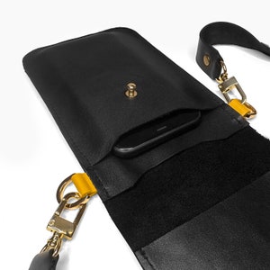 May include: Black leather crossbody phone bag with a yellow strap and a gold clasp. The bag has a flap that opens to reveal a soft black lining and a pocket for a phone.