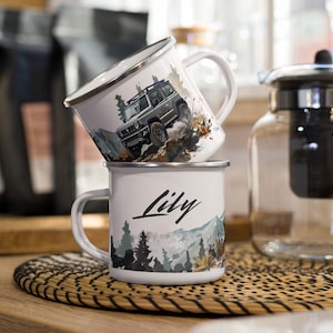 Może przedstawiać: Dwa białe kubki emaliowane ze srebrną obwódką. Górny kubek przedstawia akwarelowe przedstawienie zielonego SUV-a jadącego przez las. Dolny kubek przedstawia akwarelowe przedstawienie lasu z napisem "Lily" napisanym czarnym kursywą.