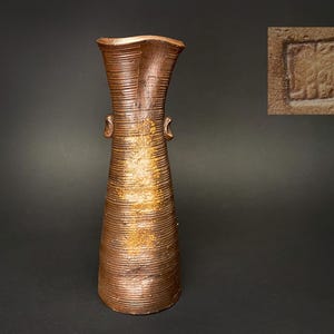 Könnte beinhalten: Eine hohe, strukturierte braune Vase mit ausgestelltem Oberteil und zwei kleinen Henkeln. Die Vase hat stellenweise eine metallisch goldene Oberfläche. Ein kleiner, quadratischer Einsatz zeigt ein gestempeltes Design.