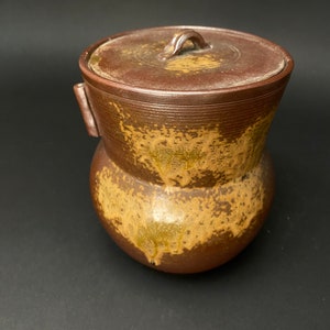 Puede incluir: Un tarro de cerámica marrón con tapa y asa. El tarro tiene un esmalte moteado con acentos marrones y dorados.