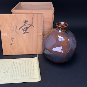 Könnte beinhalten: Eine braune Keramikvase mit glänzender Oberfläche. Die Vase ist rund und hat eine breite Öffnung. Sie steht auf einer Holzkiste mit japanischen Schriftzeichen auf dem Deckel. Die Kiste ist geöffnet und die Vase steht auf einem weißen Blatt Papier mit japanischem Text.