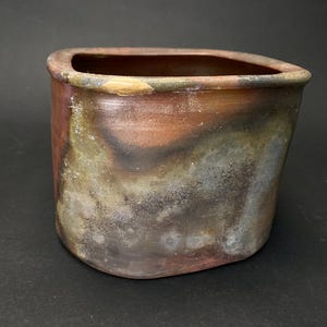 May include: A handcrafted ceramic planter with a unique, irregular shape. The exterior features a blend of earthy tones, including brown, rust, and hints of green and gray, creating a textured, organic appearance. The planter's opening is rectangular.
