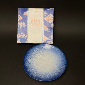May include: A blue and white ceramic plate with a raised floral design. The plate is in a box with a blue and white patterned design.