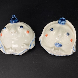 May include: Two white ceramic figurines with blue and white accents. The figurines have human-like faces with blue eyes and red cheeks. One figurine has a blue crown and the other has a blue hat.