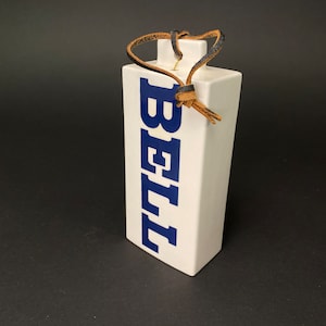 May include: White ceramic bottle stopper with a blue "BELL" logo and a brown leather strap.