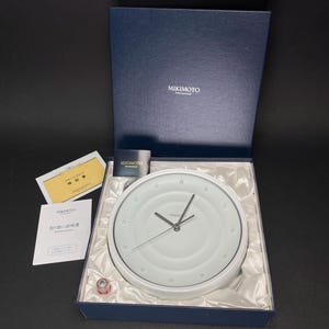 May include: A white clock with a silver face and black hands in a blue box with the word "MIKIMOTO" on the box lid. The clock is surrounded by white packaging material.