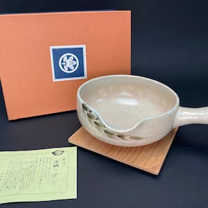Puede incluir: Un plato de servir de cerámica blanca con un asa y un diseño de hojas verdes. El plato está sobre una base de madera y viene en una caja con un logotipo japonés. La caja tiene el texto "茶碗  -  湯呑  -  お茶  -  器  -  和食器  -  日本  -  伝統  -  手作り  -  茶道  -  煎茶  -  抹茶  -  茶器  -  茶道具  -  陶芸  -  焼き物  -  うつわ  -  食器" en el lateral.
