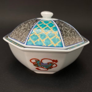 May include: A white octagonal porcelain dish with a lid. The lid has a geometric design in shades of blue, yellow, and gray. The dish has a red and blue design on the bottom.