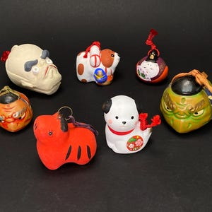 Puede incluir: Una colección de pequeñas y coloridas figuras de cerámica japonesa. Las figuras representan varios personajes, incluidos perros, demonios y otras figuras tradicionales. Las figuras están pintadas con colores brillantes y tienen detalles intrincados.