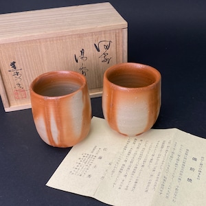 Könnte beinhalten: Zwei braune und weiße Keramik-Teetassen mit einer Holzkiste. Die Kiste trägt eine japanische Aufschrift. Die Tassen stehen auf einem Stück Papier mit japanischer Schrift.