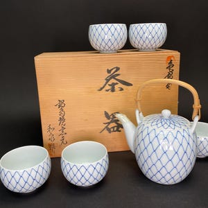 Puede incluir: Un juego de té japonés con una tetera blanca y tazas a juego, todas adornadas con un patrón de celosía azul. El juego incluye una caja de madera con caracteres japoneses. La tetera tiene un asa de bambú.