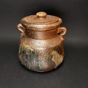 Könnte beinhalten: Ein handgefertigter Keramiktopf mit Deckel. Der Topf hat eine warme braune Farbe mit dunkleren Akzenten und einer strukturierten Oberfläche. Er hat zwei kleine Griffe und einen kleinen Knopf auf dem Deckel. Der Topf wird wahrscheinlich zur Aufbewahrung verwendet.