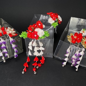 Peut inclure: Trois accessoires pour cheveux japonais, chacun dans une boîte transparente. Les ornements capillaires présentent des fleurs en tissu rouge, blanc, or et vert, avec des pétales en tissu blanc, rouge et violet pendants. Des cloches argentées sont aux extrémités.